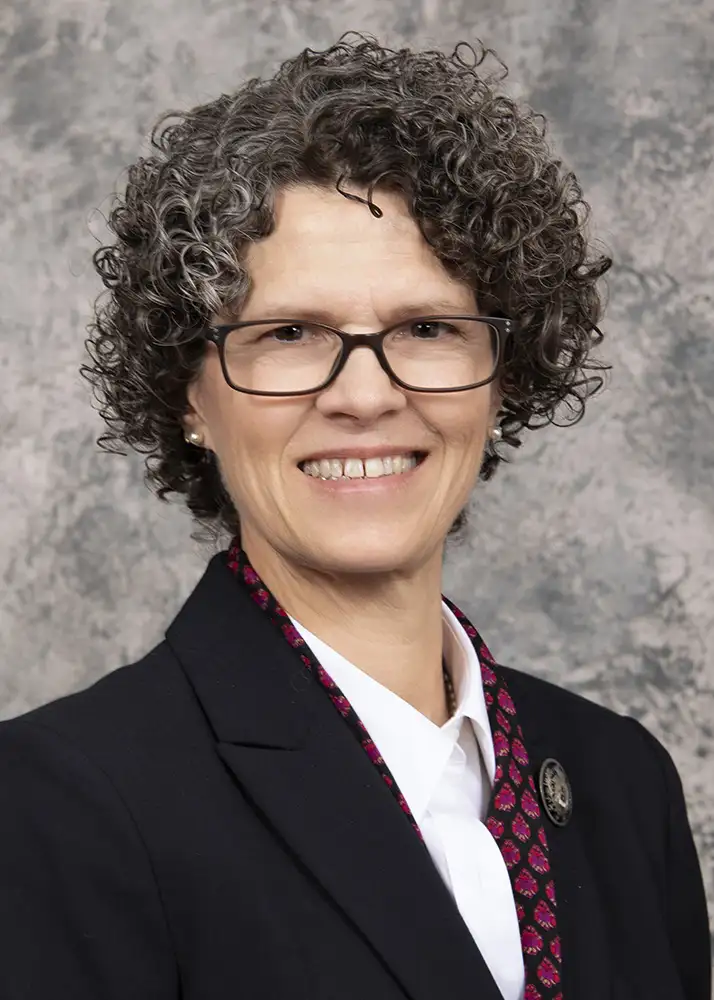 Woman in professional clothing with curly hair and glasses, smiling at the camera